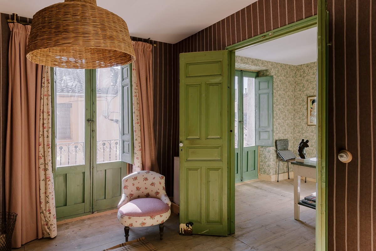 Dormitorio de estilo rústico con muebles de madera, papel pintado de flores. Decoración en tonos verdes, lámpara de mimbre y cortinas. Ambiente cálido y ecléctico.