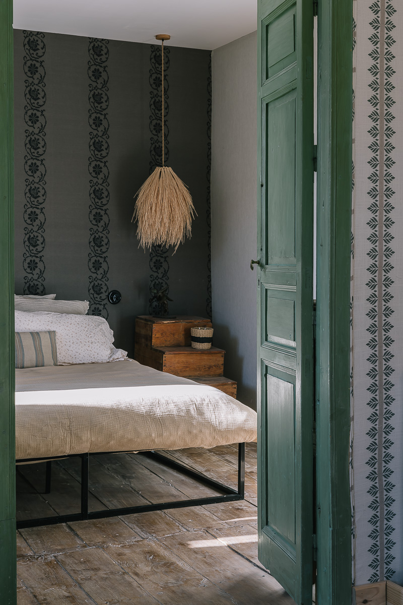 Dormitorio de estilo rústico con muebles de madera, papel pintado de flores. Decoración en tonos verdes, lámpara vintage. Ambiente cálido y ecléctico. Materiales orgánicos.